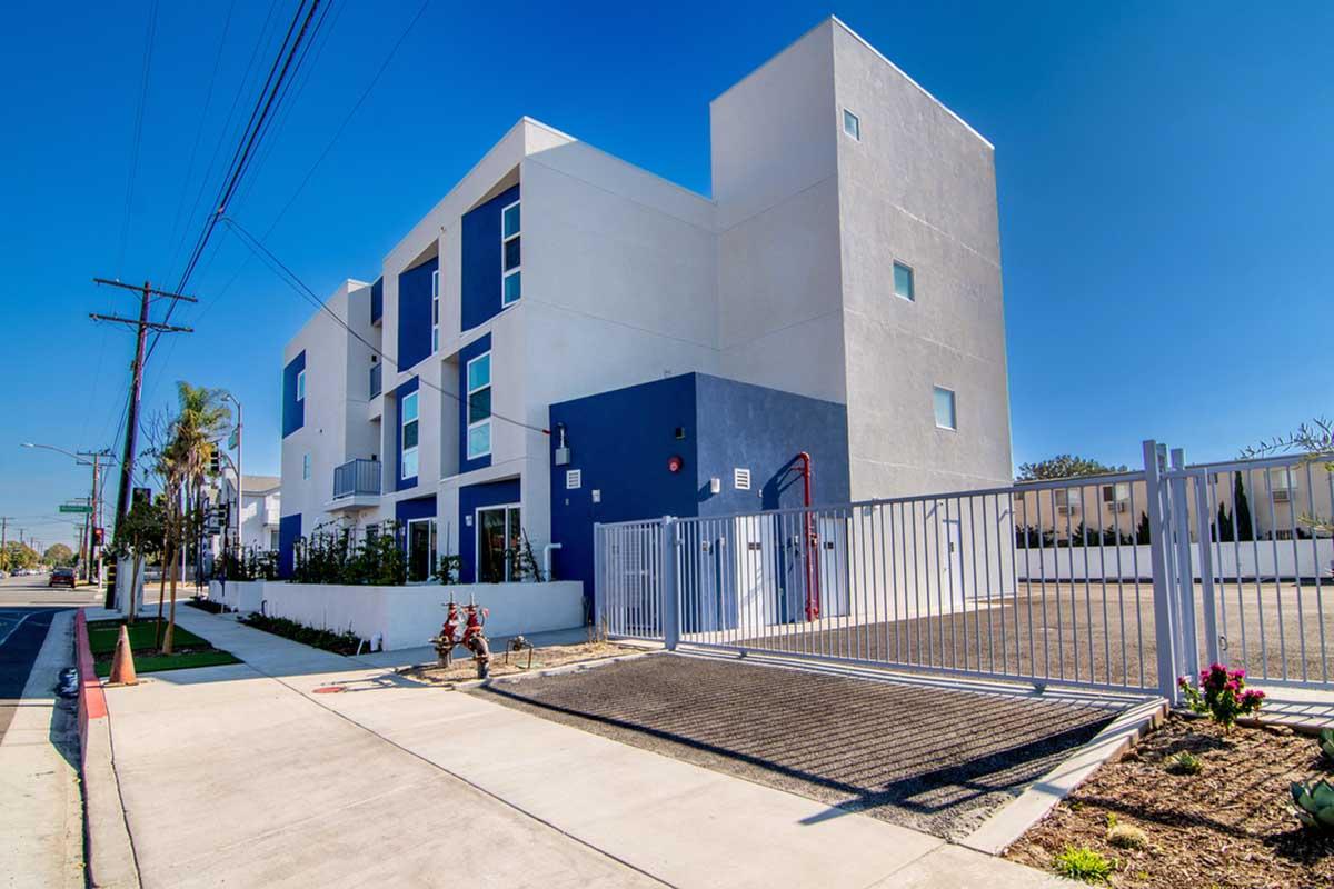 Modern building with a mix of blue and white exterior, featuring large windows. The structure has three stories and is surrounded by greenery, including palm trees. A gated entrance is visible in front, and the scene is set on a sunny day with clear blue skies. Sidewalks and utility poles line the street.