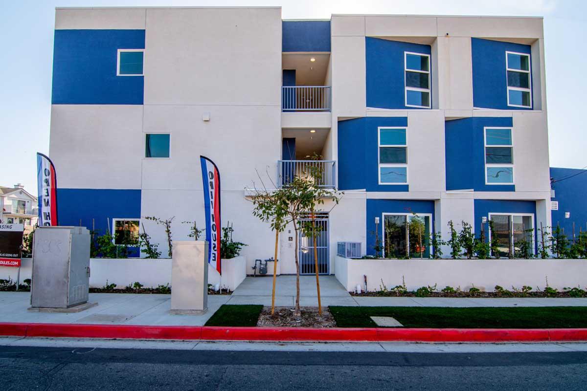 A modern three-story residential building with a mix of white and blue exterior. The front features glass windows, a small tree, and landscaped areas. Two flags are displayed outside, and the street is visible in front of the building, showcasing a clean and contemporary architectural design.