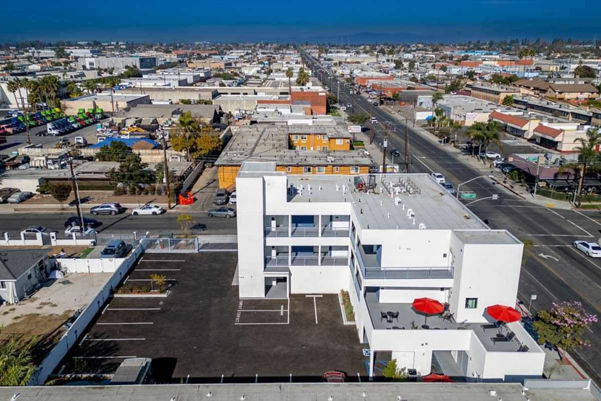 Aerial view of a modern, white multi-story building with a flat roof and red umbrellas on a paved parking lot. Surrounding urban area features various buildings and roads, with a clear blue sky above. The scene captures a mix of residential and commercial properties in a bustling neighborhood.
