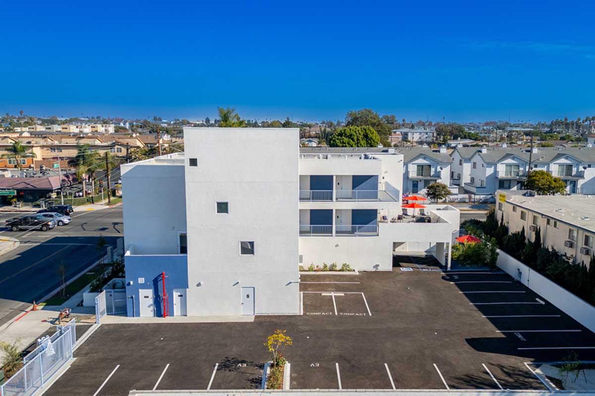 A modern white multi-story building with balconies and a flat roof, situated in a suburban area. The surrounding landscape includes residential homes, palm trees, and clear blue skies. A parking lot in front has several marked spaces, and the street is visible alongside the property.