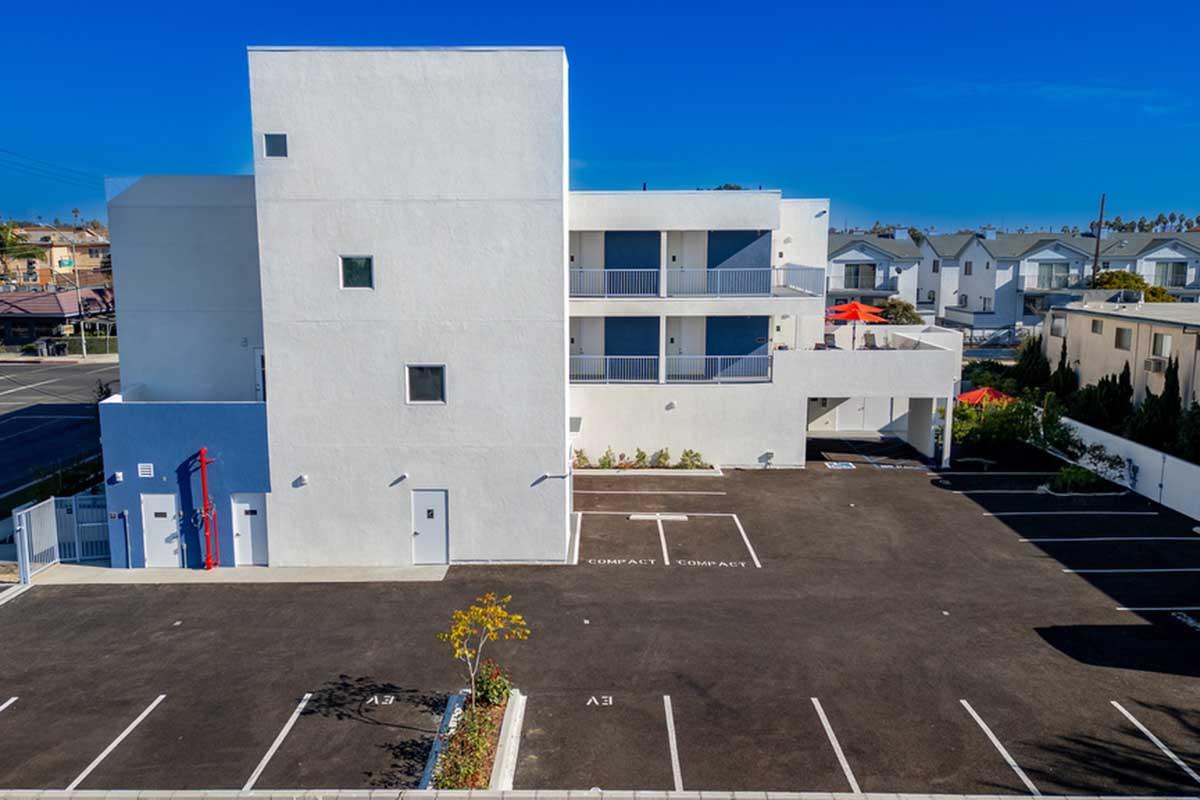 Modern white building with multiple windows and balconies, set against a clear blue sky. The foreground features a paved parking lot with designated compact car spaces, surrounded by greenery. Nearby structures in the background suggest a residential area.