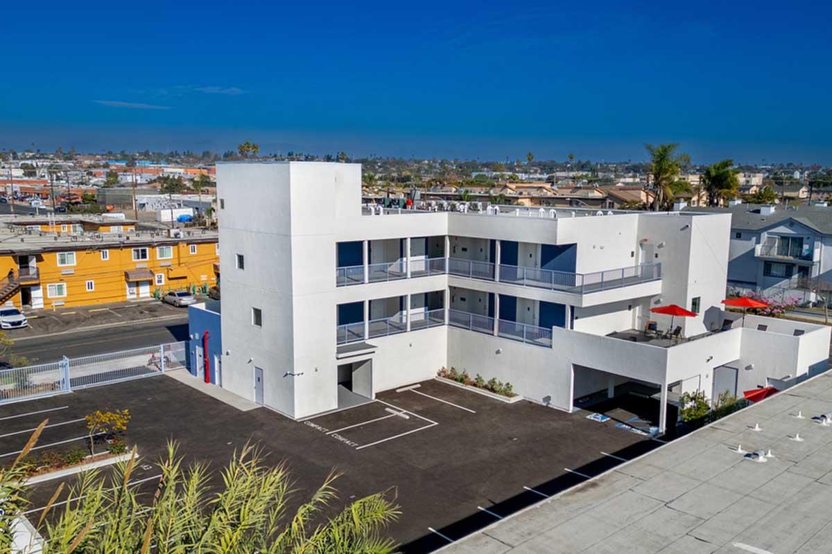 A modern, two-story white building with multiple balconies and a clean parking area. The structure is surrounded by greenery and the urban landscape includes colorful buildings in the background under a clear blue sky.