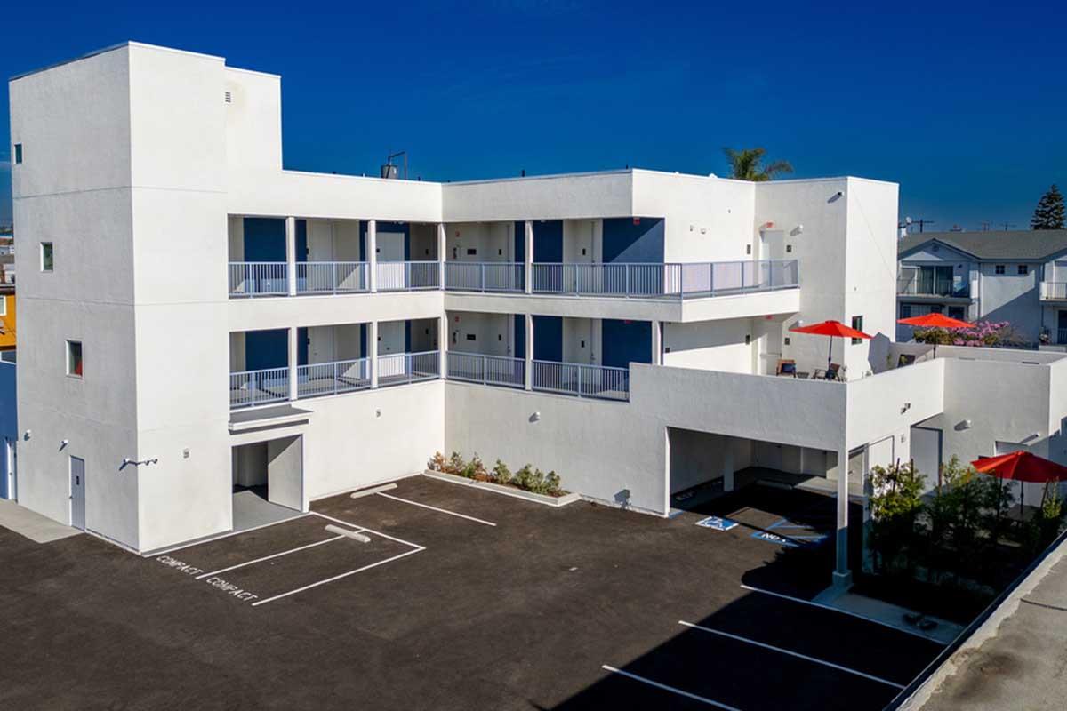 A modern, multi-story white building featuring balconies, surrounded by paved parking spaces. Bright blue skies are in the background, and there are red umbrellas on the upper level. The structure has a clean, contemporary design with landscaping near the entrance.