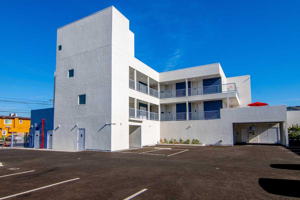 A modern, three-story white building with multiple balconies. The structure features a flat roof and large windows. In front, there's a parking lot with empty spaces. A red umbrella is visible on the upper floor, and the sky above is clear and blue. The overall setting appears well-maintained and inviting.
