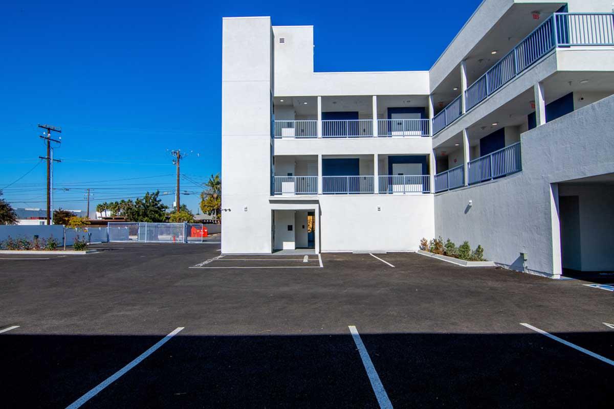 A modern, multi-story building featuring white exterior walls, balconies, and an asphalt parking lot. The scene is under a clear blue sky, with parked vehicles visible in marked spaces. The surrounding area includes some plants and power lines in the background.