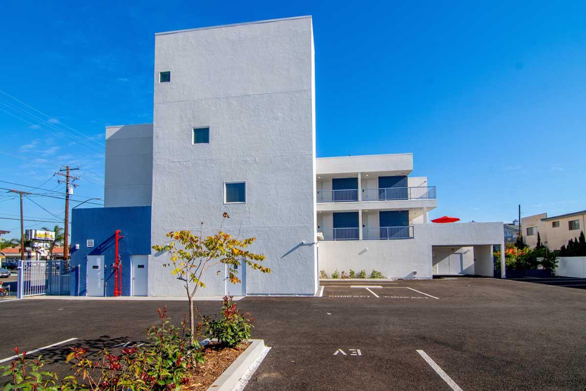 Modern white building with multiple stories, featuring large windows and a balcony. The surrounding area includes a paved parking lot with marked spaces, some greenery, and a clear blue sky. Power lines are visible in the background, contributing to an urban atmosphere.