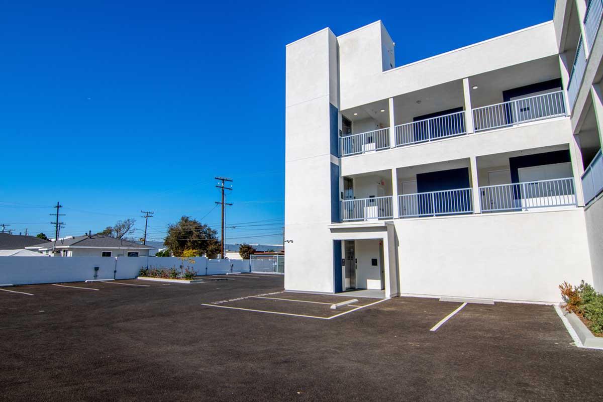 A modern, white multi-story building with balconies, set against a clear blue sky. The foreground features a paved parking lot with marked spaces, and a few trees are visible in the background. The scene conveys a bright and clean urban environment.