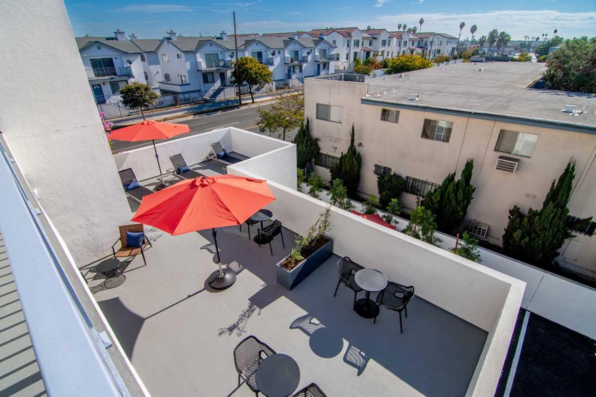 A rooftop terrace featuring blue skies and several orange umbrellas. Below, there are tables and chairs arranged for seating. Surrounding buildings are partially visible, along with palm trees in the background. The scene depicts a sunny, inviting outdoor space perfect for relaxation or socializing.