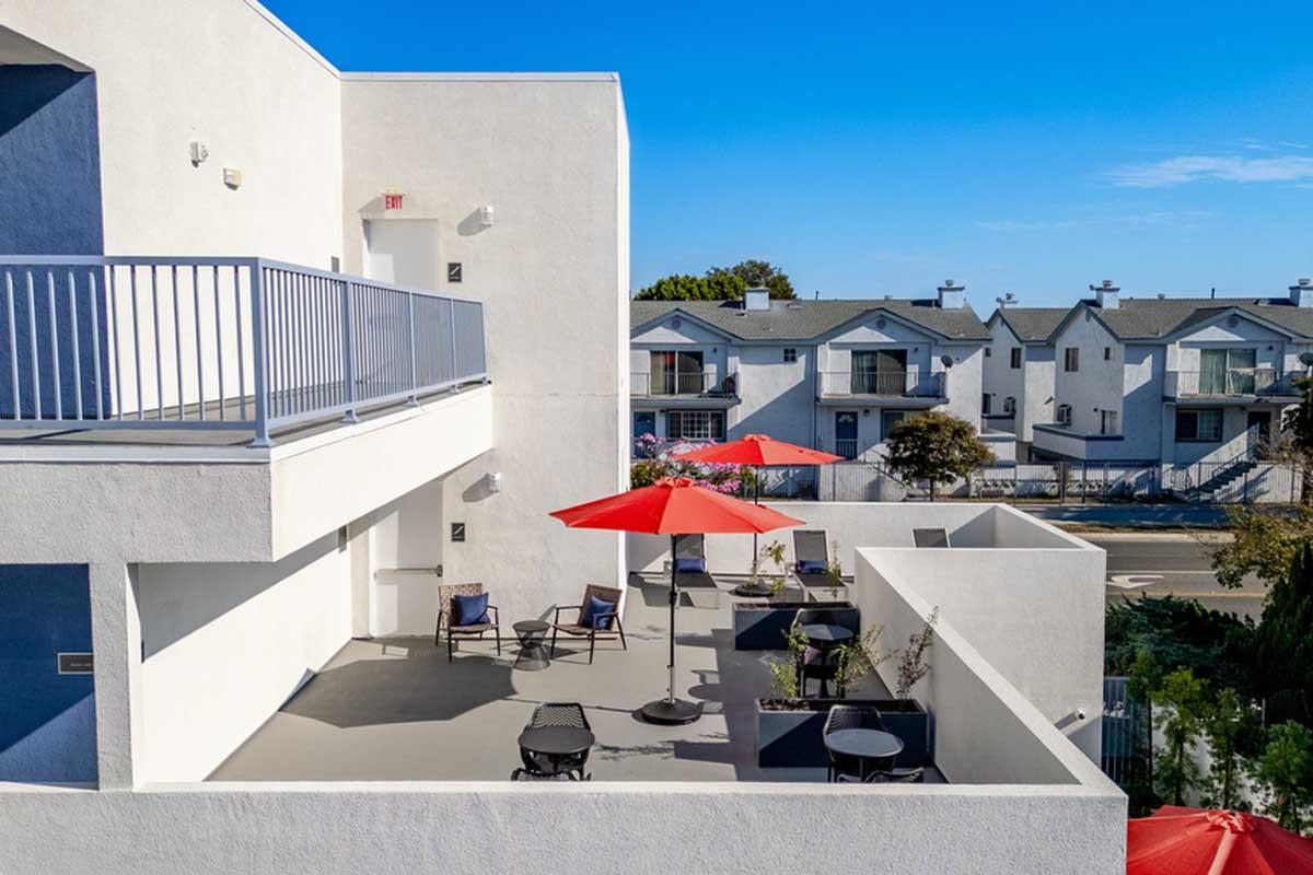 A sunny outdoor terrace featuring several seating areas with black chairs and red umbrellas. In the background, a row of white buildings with multiple rooftops is visible under a clear blue sky. The scene conveys a relaxed and inviting atmosphere.