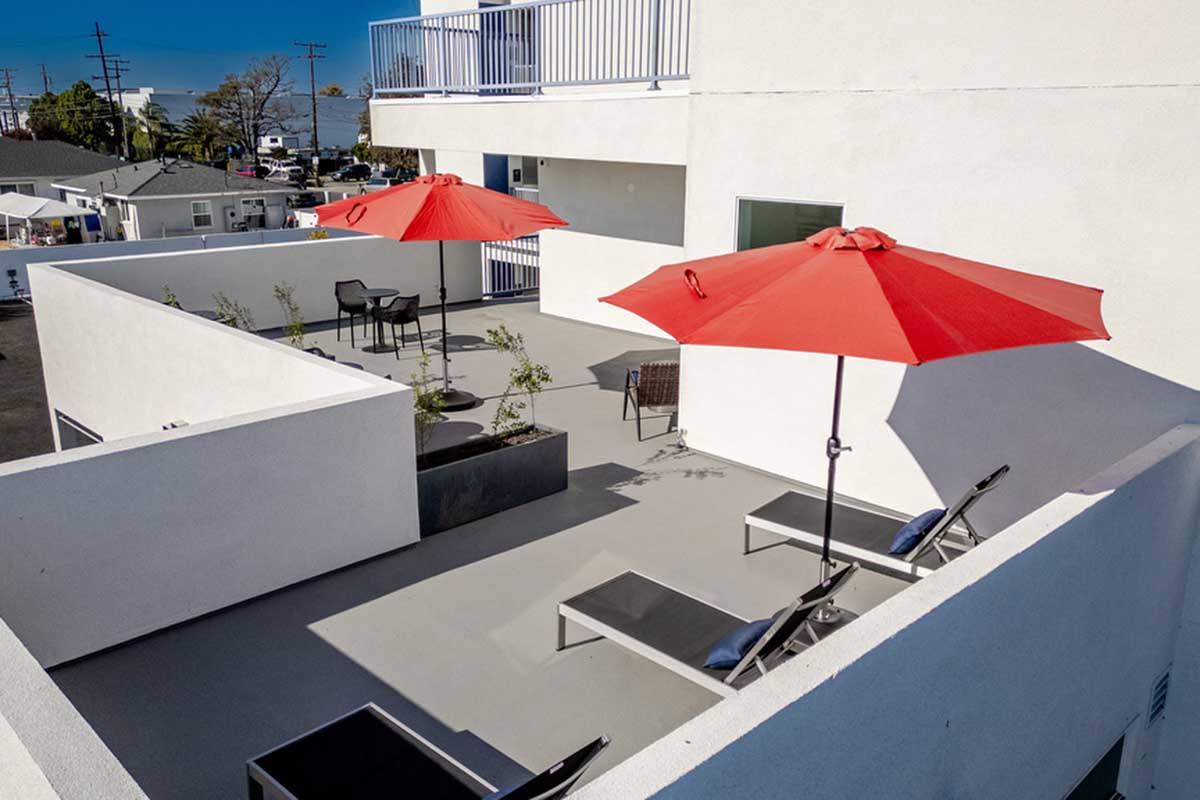 A modern rooftop terrace featuring two large red umbrellas over lounge chairs, surrounded by minimalist landscaping. The space is designed for relaxation, with a seating area and a clear sky above, providing a view of nearby buildings and the landscape beyond.