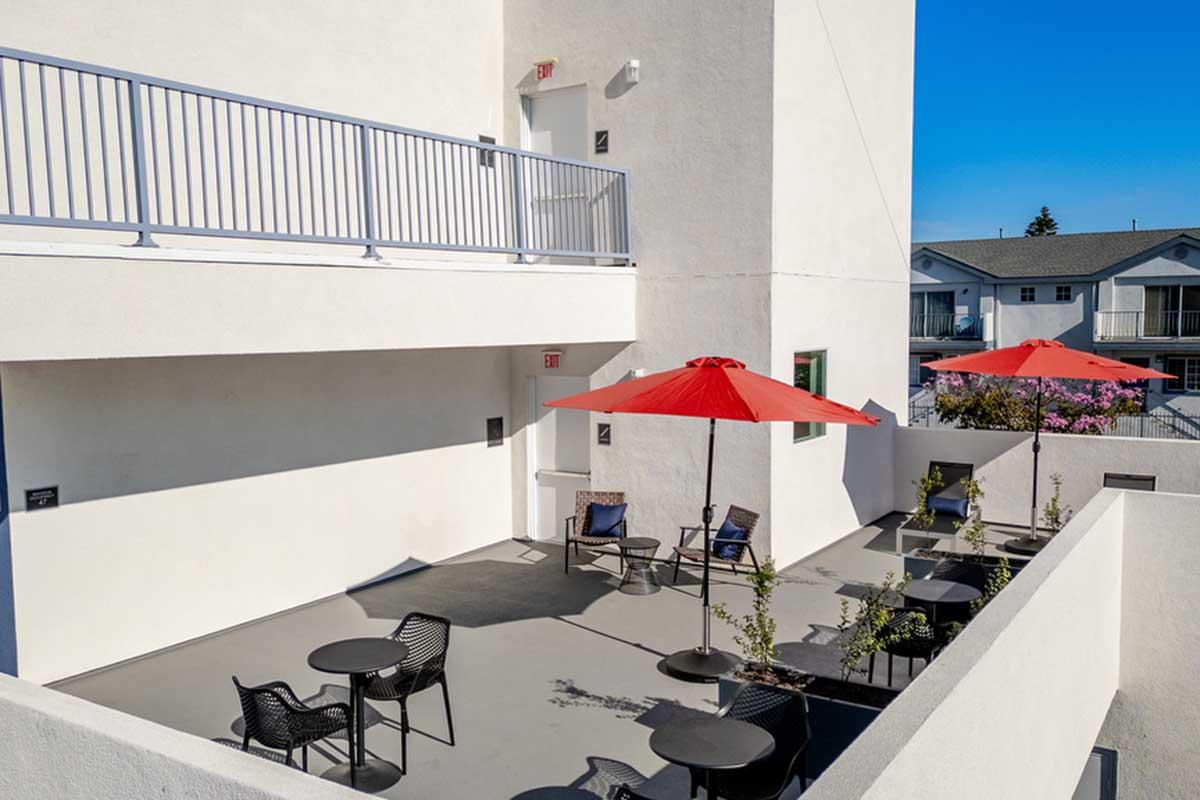 A bright outdoor seating area with several black chairs and small tables, shaded by red umbrellas. The wall is white, and there is a balcony above with a railing. Bright blue sky in the background, with some nearby buildings visible.