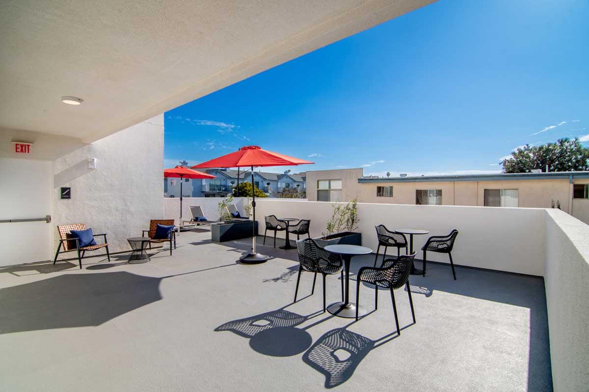 A rooftop terrace featuring black outdoor seating and tables, shaded by red umbrellas. The surrounding area includes a few potted plants and a clear blue sky, adding to the inviting atmosphere of the space.