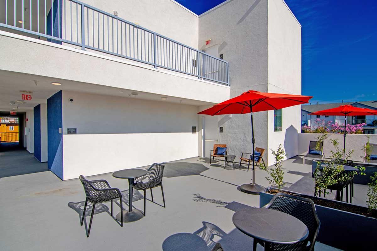 A modern outdoor space featuring a white building with a balcony, surrounded by vibrant flowers. There are several black patio tables and chairs, along with red umbrellas providing shade. The clear blue sky offers a bright backdrop, creating a welcoming atmosphere for relaxation or socializing.