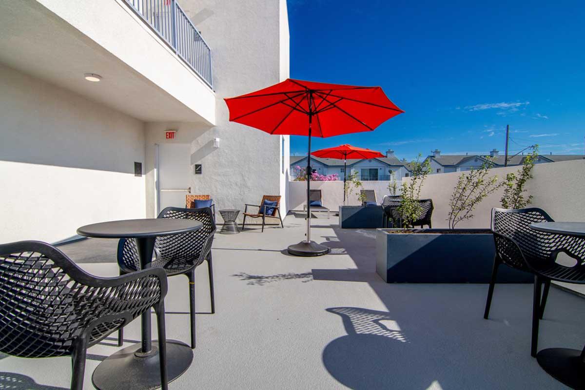 Outdoor terrace with modern black chairs and tables under bright red umbrellas, surrounded by potted plants. The bright blue sky is visible above, creating a vibrant and inviting atmosphere for relaxation.