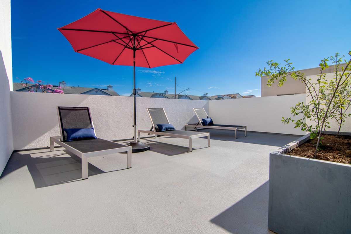 A sunlit terrace featuring two sleek lounge chairs with dark cushions under a large red umbrella. A potted plant is seen in the foreground, and a clear blue sky forms a bright backdrop, enhancing the relaxing atmosphere of the space.