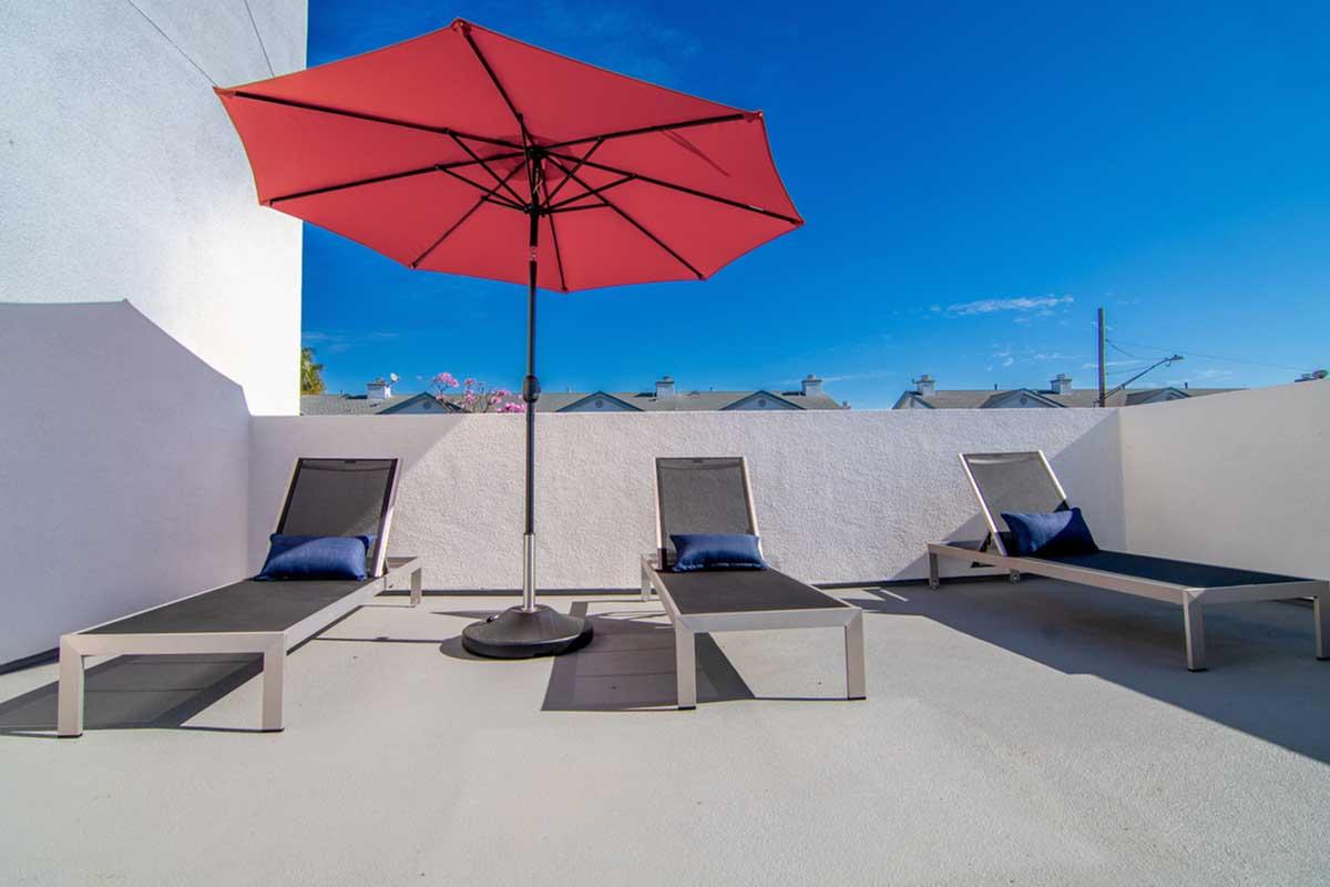 A sunlit outdoor space featuring three modern lounge chairs with dark fabric, positioned under a bright red umbrella. The setting includes a smooth concrete floor and a clear blue sky in the background. Some residential rooftops are visible in the distance.