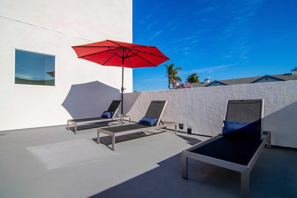 A serene outdoor patio with two modern lounge chairs and a large red umbrella providing shade, set against a clear blue sky. A white wall and a nearby window complement the minimalist design, creating a relaxing space ideal for sunbathing or enjoying a quiet moment.