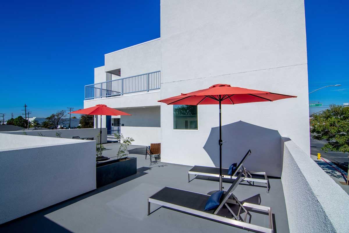 A rooftop terrace featuring two lounge chairs and red umbrellas. The space has a modern design with white walls and a clear blue sky in the background. Plants are placed around the terrace, adding greenery to the urban setting.