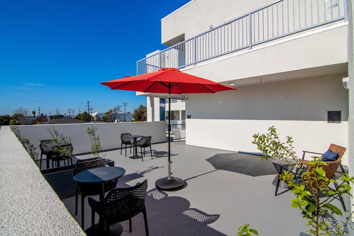 A rooftop terrace featuring several outdoor seating arrangements, including black chairs and tables, with a large red umbrella providing shade. The area is bathed in sunlight under a clear blue sky, and there are small plants in the corners, enhancing the inviting atmosphere.