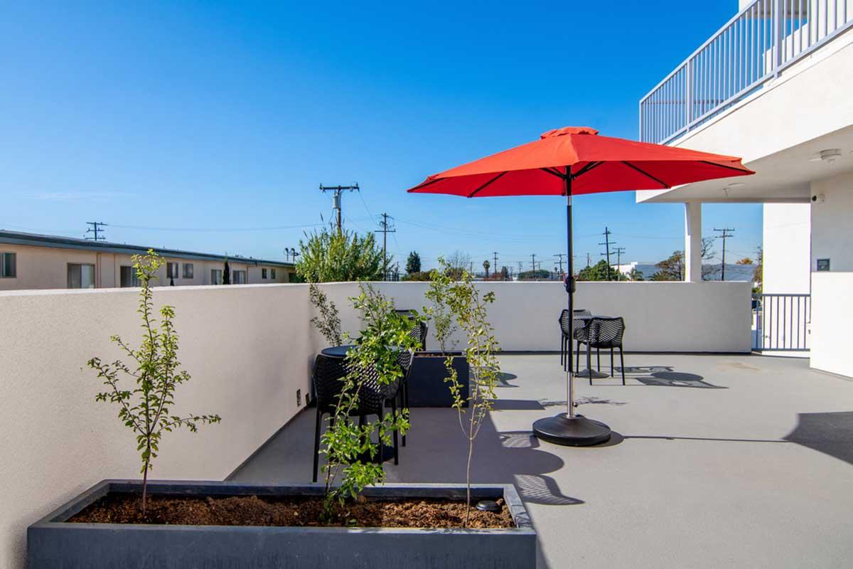 A bright outdoor patio featuring a red umbrella over a black seating area, surrounded by small plants in planters. The sky is clear and blue, with a few buildings and power lines visible in the background.