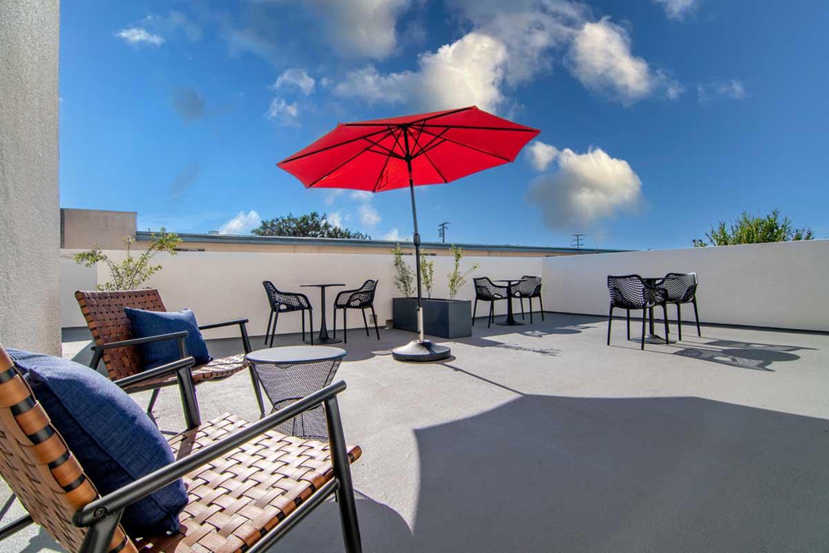 A rooftop terrace featuring several black chairs and tables, a large red umbrella providing shade, and blue sky with a few clouds in the background. The space is designed for relaxation, with a clean, modern aesthetic.