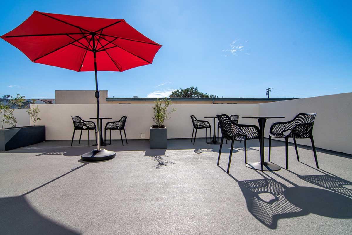 A rooftop terrace featuring several black tables and chairs under a vibrant red umbrella. The space is bright and sunny, with a clear blue sky and a few small potted plants adding greenery to the modern setting. Shadows are cast by the furniture on the concrete surface.