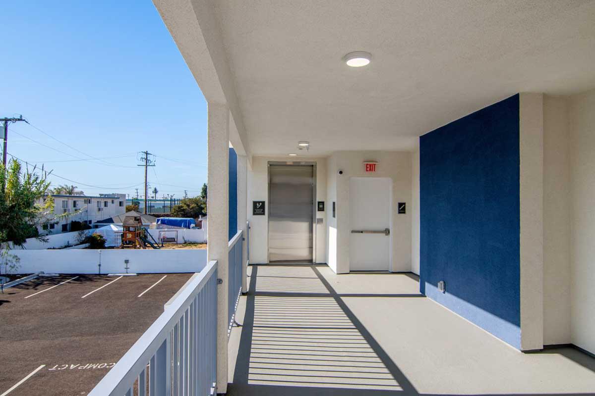 A well-lit corridor featuring an elevator and two doors leading to rooms, with a blue accent wall. The scene includes a view of a parking lot and other buildings in the background, indicating an outdoor environment. Shadow lines from the railing create a geometric pattern on the floor.