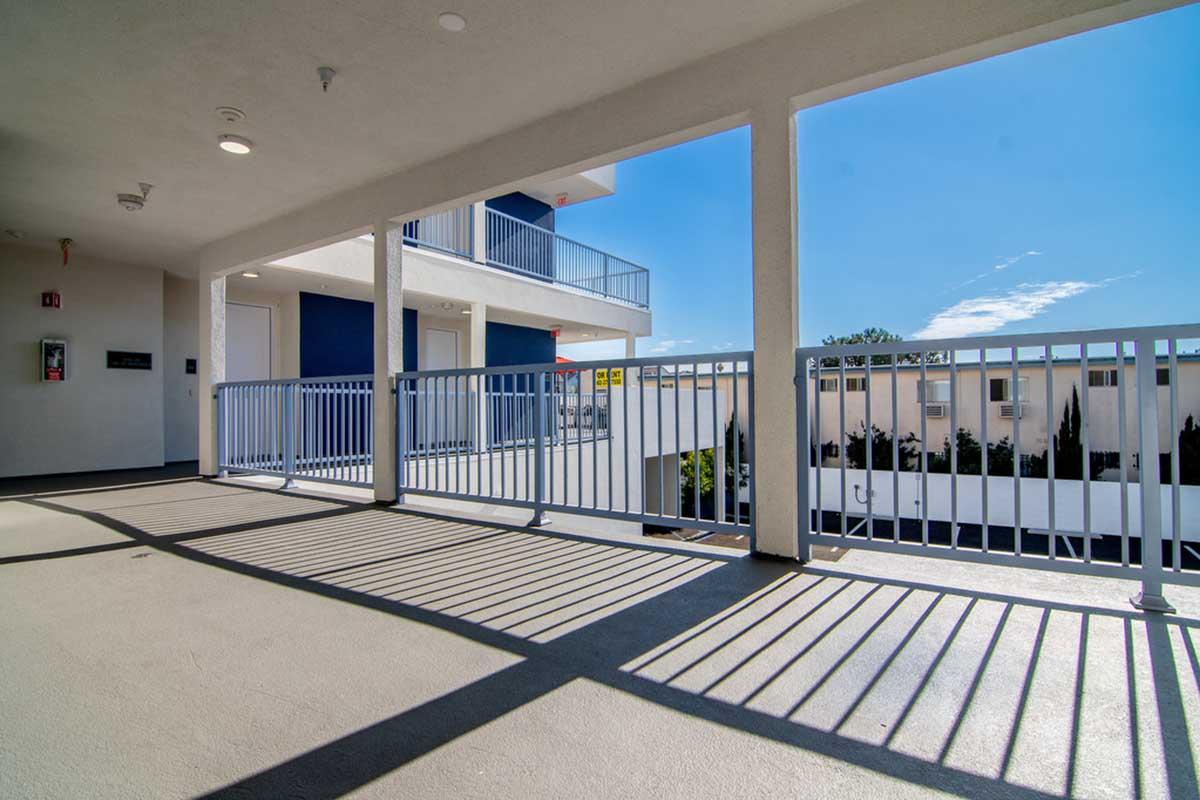 A view of a modern balcony or corridor within a building. The space features railings casting soft shadows on the floor, with blue skies and other buildings visible in the background. Natural light illuminates the area, creating a bright and open atmosphere.
