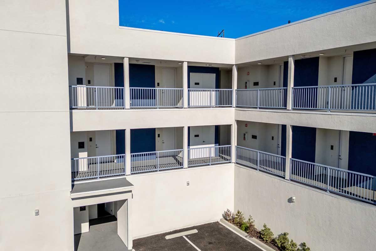 Two-story building with a modern exterior featuring white walls and blue doors. The balcony area is visible, showing railings and multiple doorways. The ground floor has an entrance leading to a parking area with a few small plants along the building's base. Clear blue sky in the background.