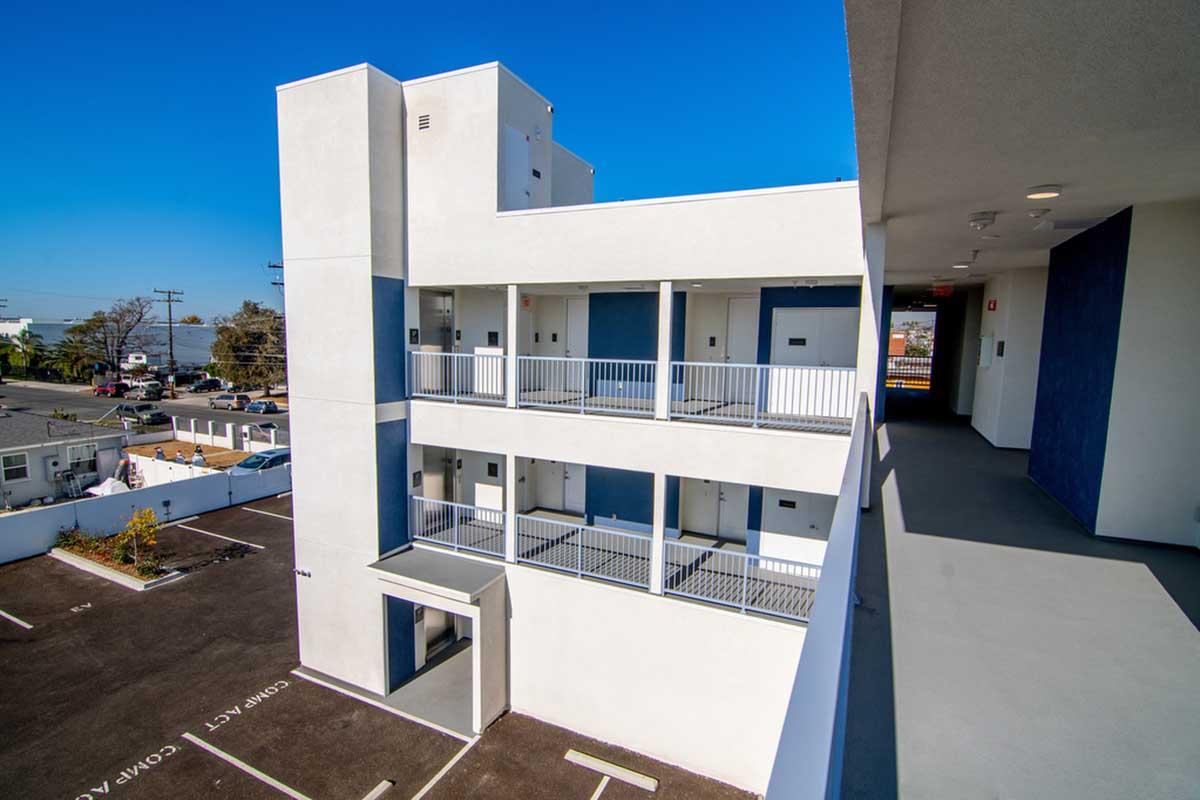 Newly constructed multi-story building with white exterior and blue accents. Balconies are visible on each floor. The ground level includes a parking area with marked spaces. Clear blue sky in the background, giving a bright and sunny atmosphere.