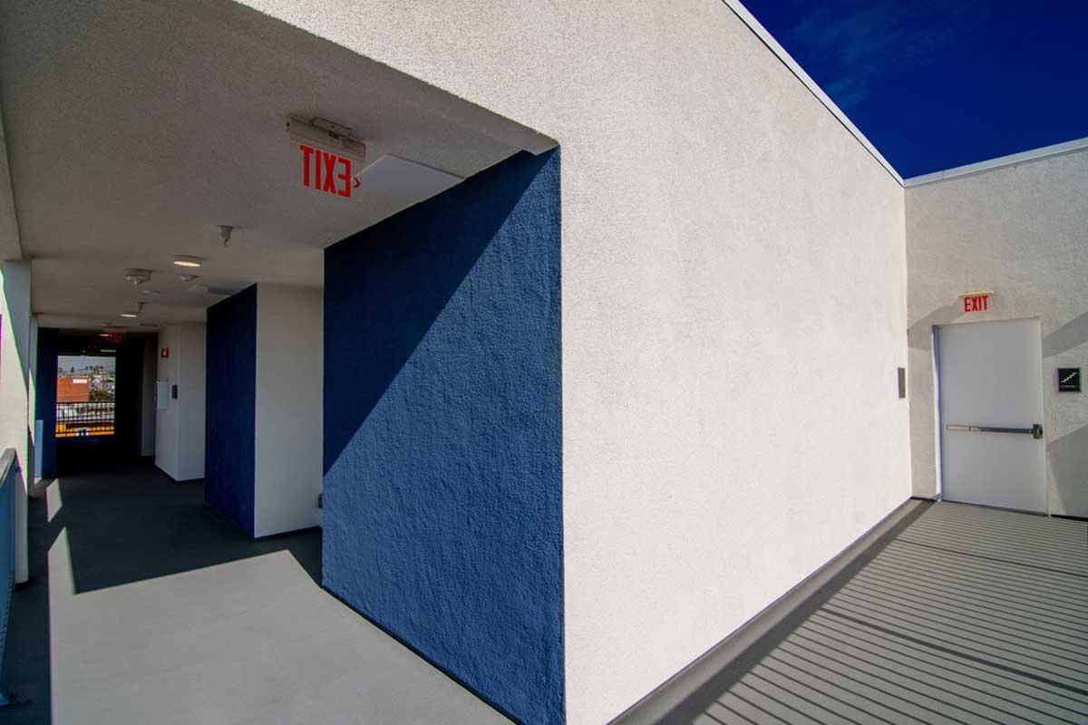A modern hallway with a white textured wall and a blue accent wall. The area is well-lit with bright overhead lights, and the floor is gray. Visible exit signs indicate emergency exits. The perspective is from a corner, showing a glimpse of the adjacent hallway and doorways. The sky is clear and blue outside.