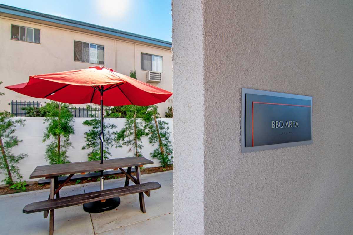 A cozy BBQ area featuring a wooden picnic table under a red umbrella, surrounded by greenery and a white wall. A sign indicating "BBQ AREA" is mounted on a nearby wall. The setting is bright and inviting, perfect for outdoor gatherings.