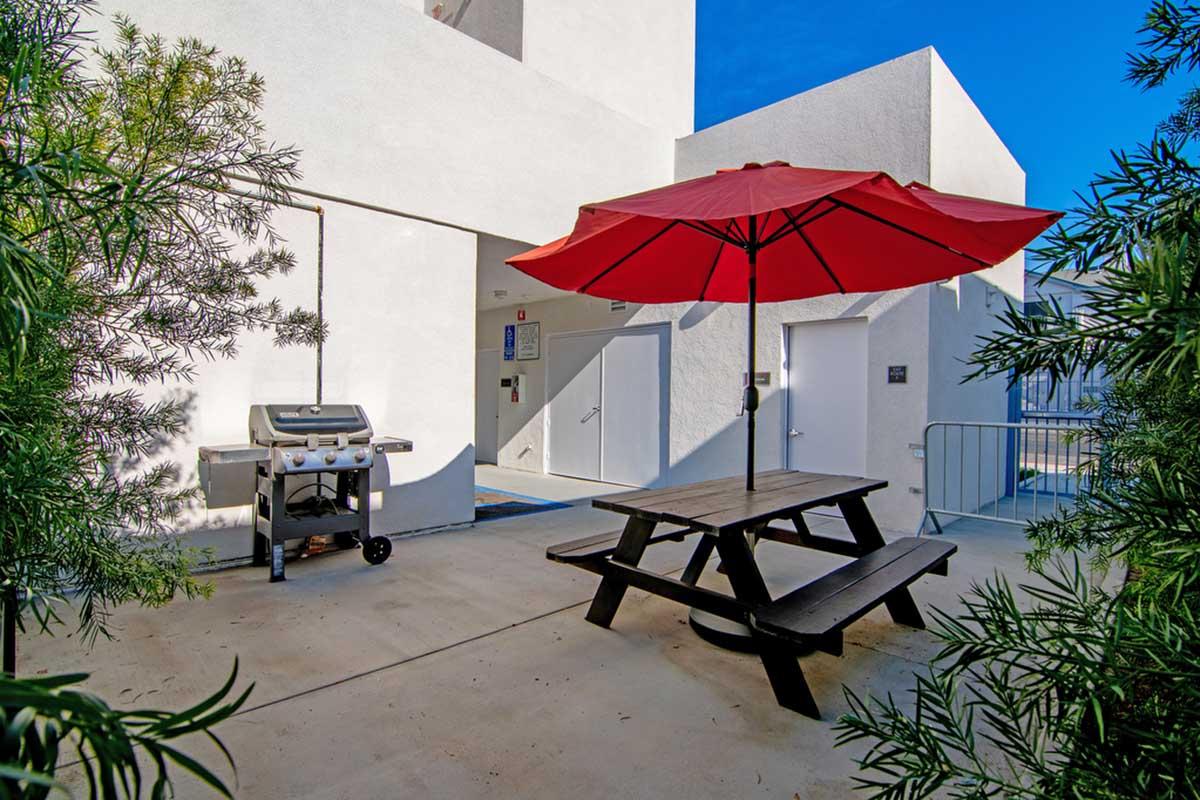 A shaded picnic area featuring a wooden table with a red umbrella. Nearby, a grill is positioned against a white wall, with a clear blue sky above. Lush green foliage frames the scene, creating a welcoming outdoor space.