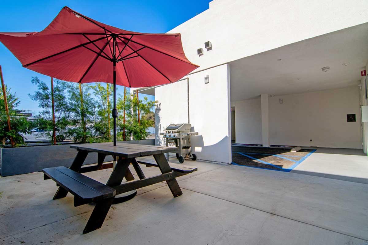 A patio area featuring a black picnic table with a red umbrella, a barbecue grill nearby, and a clean concrete surface. Lush greenery is visible in the background, with a bright blue sky overhead. The scene suggests a sunny outdoor space ideal for relaxation and gatherings.