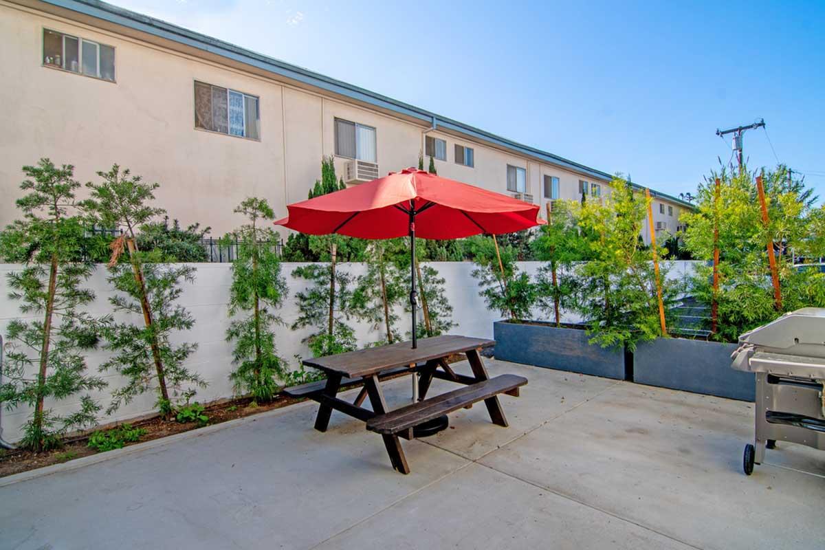 A cozy outdoor patio featuring a wooden picnic table with an red umbrella, surrounded by green shrubs. In the background, a low white wall encloses the area, and a grill is visible nearby. The setting is bright and inviting, ideal for dining or relaxation.