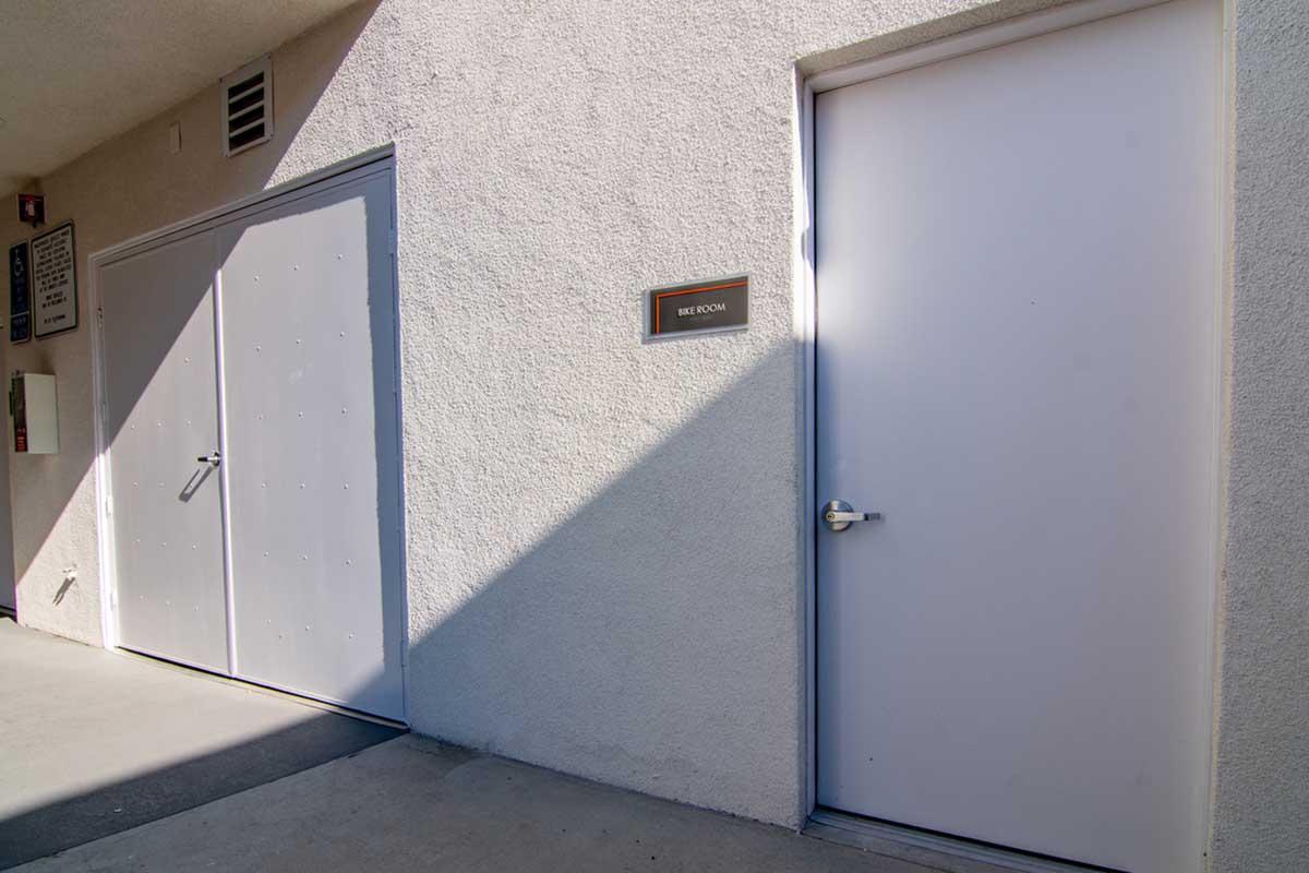 A hallway with two doors: one gray door on the left with handles and a sign above, and a white door on the right. The area is well-lit, with sunlight casting shadows on the wall. The surface is plain and unadorned, creating a minimalist aesthetic.