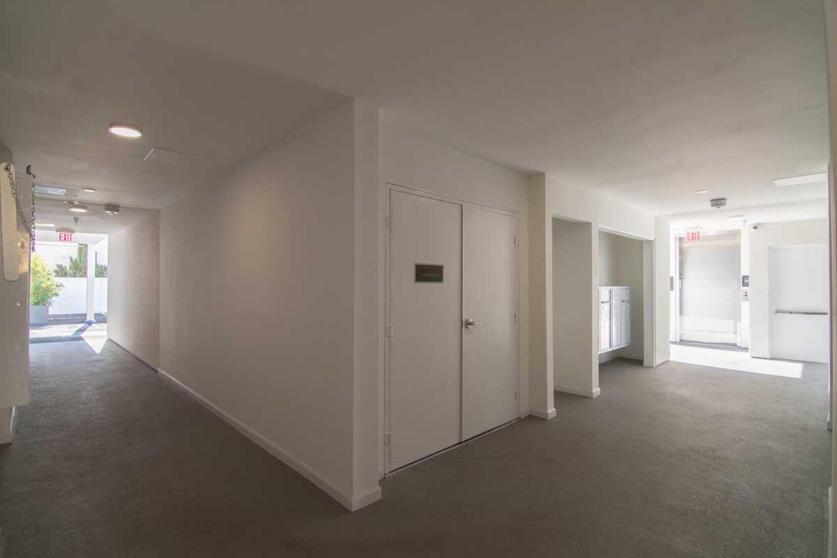 A modern hallway with white-painted walls and a light gray carpet. The corridor features a closed door with a sign, leading to a bright area with natural light. An elevator is visible at the end of the hallway, along with a small kitchenette or utility area on the right. The space appears clean and well-maintained.