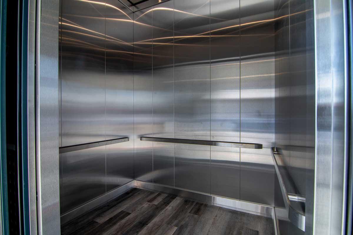 Interior view of a modern elevator featuring sleek stainless steel walls, a clean floor, and horizontal lighting. The elevator is empty, showcasing two handrails along the sides.