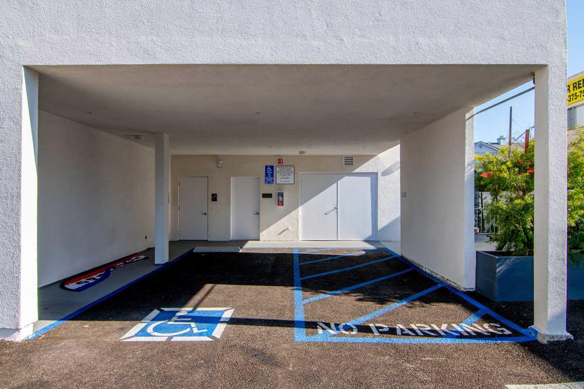 A vacant parking area beneath a building, featuring a designated handicap parking space marked with a blue symbol, and a "No Parking" sign painted on the ground. The walls are plain white, and there are entries visible for two doors. The area is well-lit and tidy.