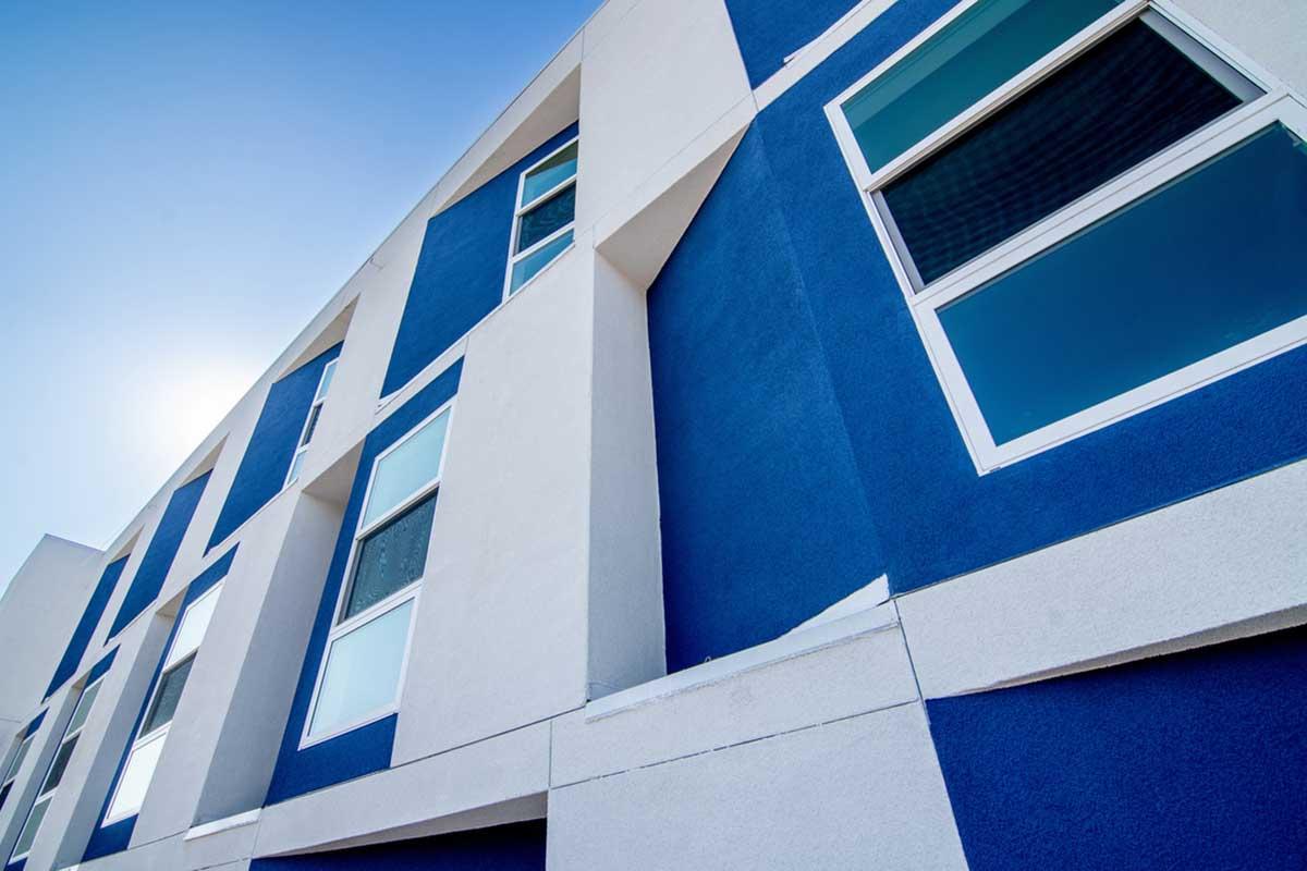 A close-up view of a modern building with a geometric design, featuring large windows and a blue and white color scheme. The angle captures the facade's texture and the clear blue sky in the background, emphasizing the contemporary architectural style.
