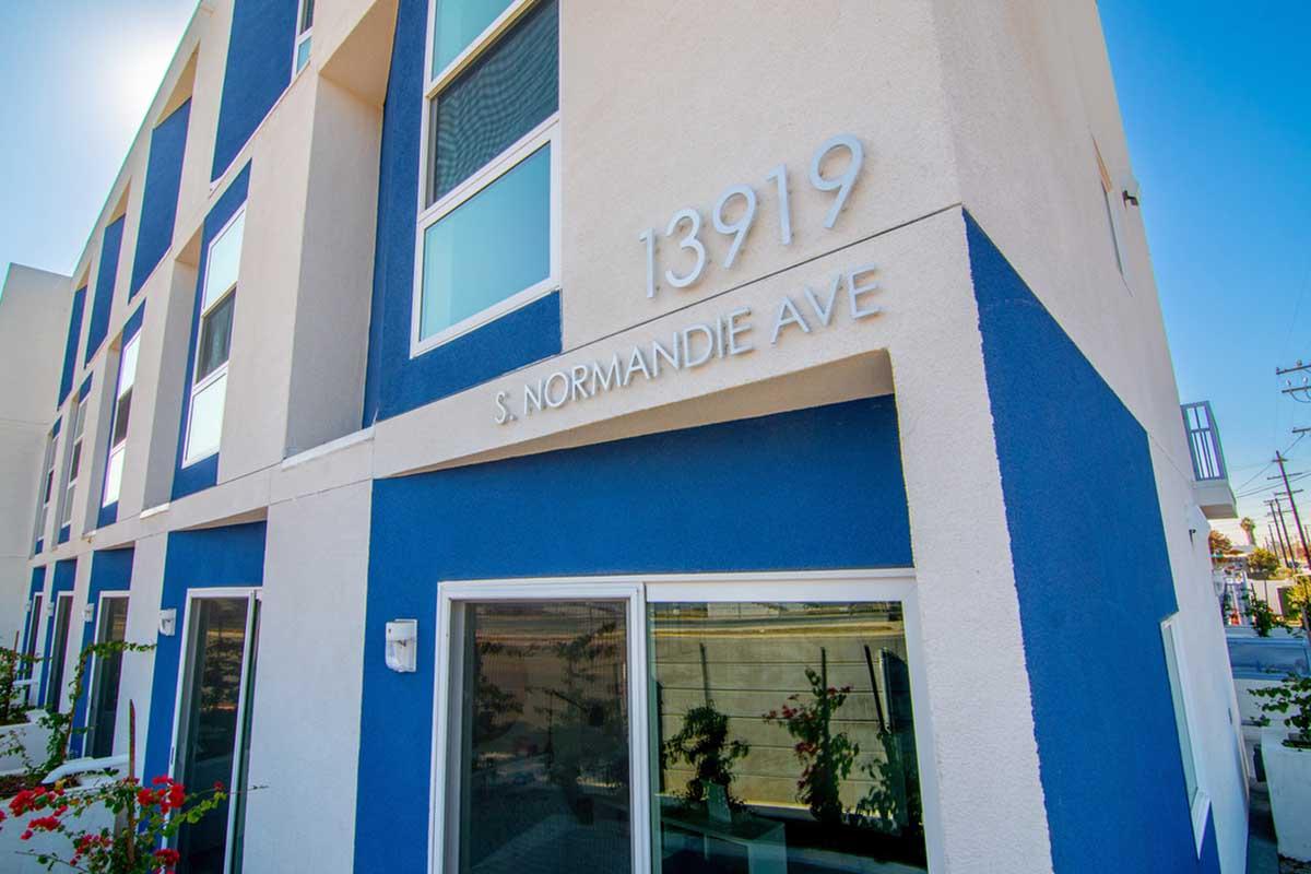 Building exterior featuring a modern design with a prominent sign displaying "13919 S. Normandie Ave." The structure has a mix of white and blue colors, large windows, and nearby flowering plants. The scene is well-lit, suggesting a sunny day.