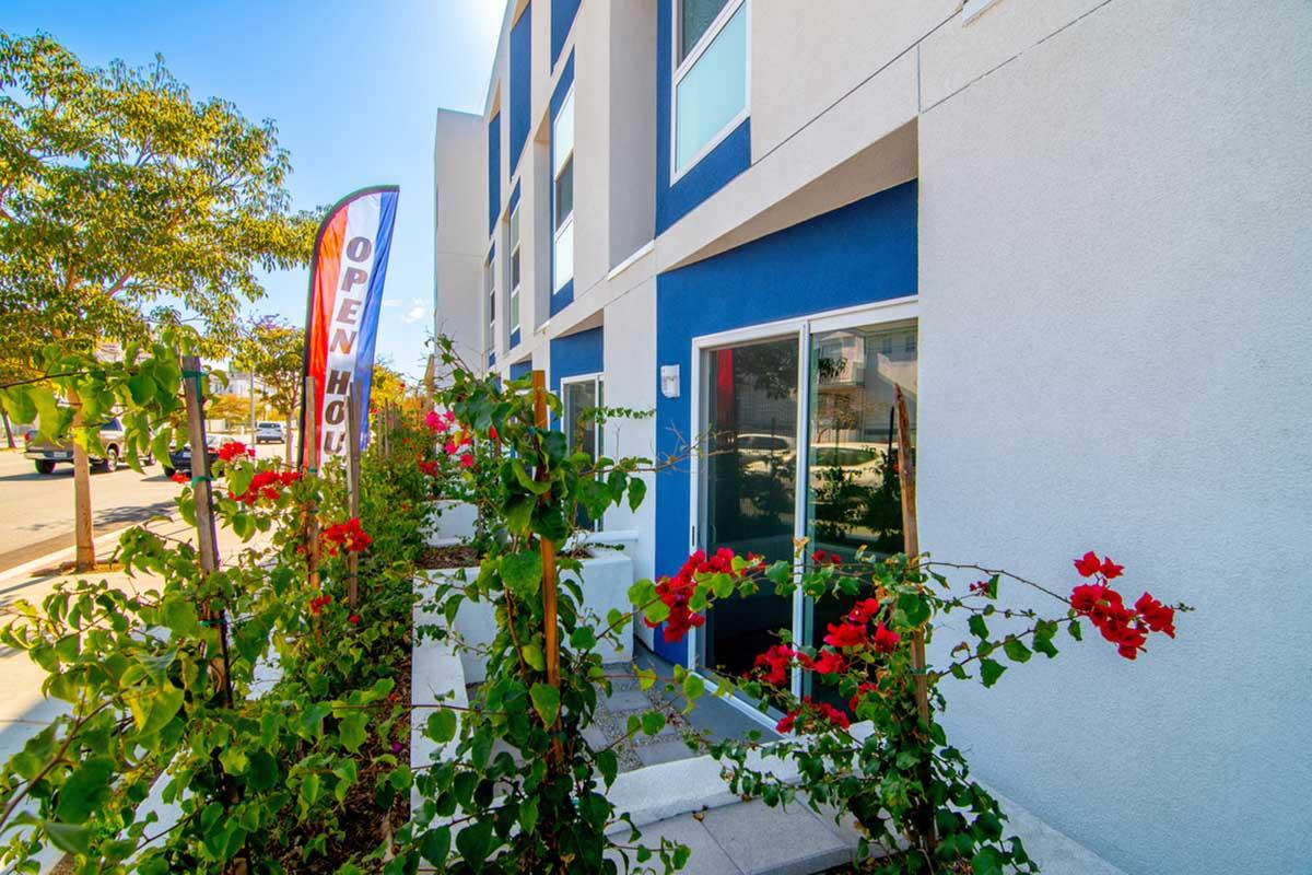 Exterior view of a modern building with a blue accent wall, featuring a colorful "OPEN HOUSE" banner. In the foreground, there are vibrant red bougainvillea flowers growing along the pathway, which is bordered with greenery. The sky is bright and clear, enhancing the welcoming atmosphere.
