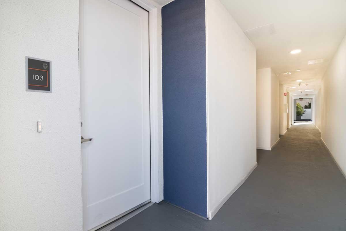 A hallway with a closed white door marked "103" on the left. The walls are painted white with one side featuring a blue accent wall. The flooring is gray, and there are illuminated pathways leading further down the hallway, with a view of more doors and a window at the end.