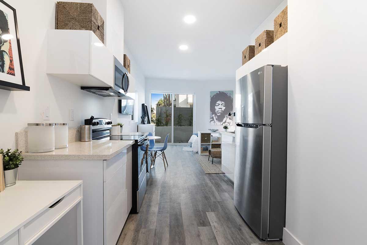 Modern kitchen and living area with sleek white cabinetry, stainless steel appliances, and a wooden floor. Natural light floods the space through a large window. Decorative storage boxes are on top of the cabinets, and a minimalist dining table is visible in the foreground.