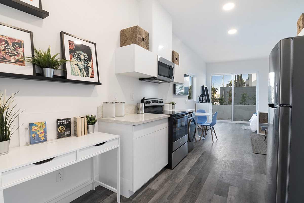 Modern kitchen with white cabinetry, stainless steel appliances including an oven and refrigerator, and a small dining area with blue chairs. Decorated with framed art on the walls and plants, the space has a bright, airy feel with natural light coming in from the window.