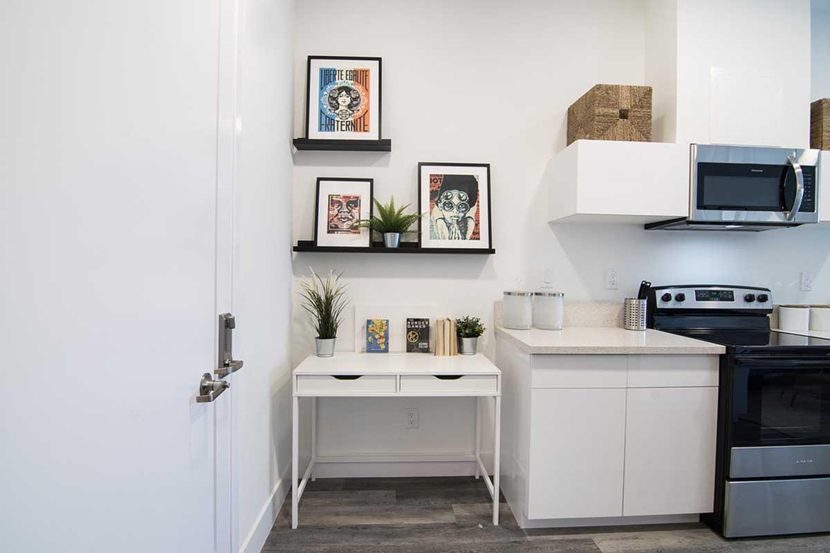 A modern kitchen corner featuring a white desk with two drawers, decorated with books and small plants. Above the desk are two black shelves displaying framed artwork. The kitchen includes stainless steel appliances, including a microwave and stove, with a clean, minimalist design and light-colored walls.