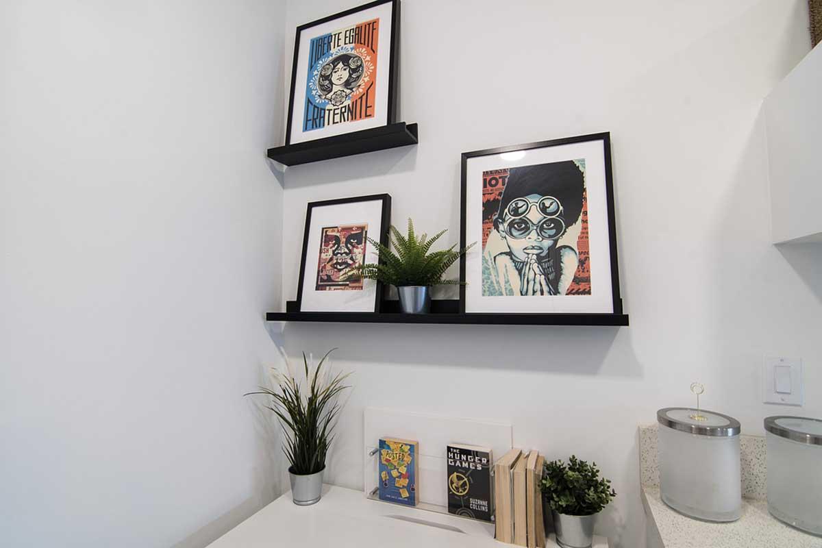 A modern corner featuring two black frames holding colorful art prints on wall-mounted shelves. Below, a small potted plant and a book stand with several books. The decor reflects a blend of artistic styles and vibrant colors, enhancing the space's aesthetic appeal.