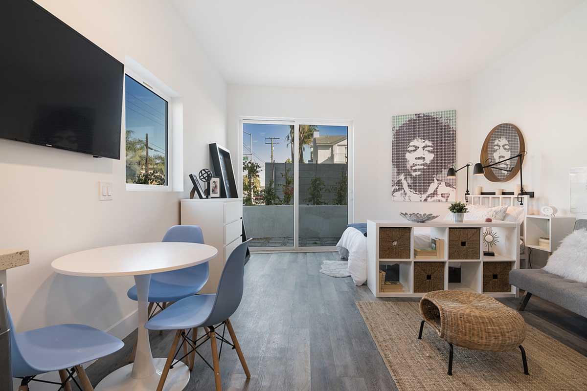 A modern, bright living space featuring a small round dining table with blue chairs, a television mounted on the wall, and a large window letting in natural light. A multi-purpose storage unit and a comfortable sofa are present, along with a decorative woven stool and a prominent wall art piece depicting a figure's portrait.
