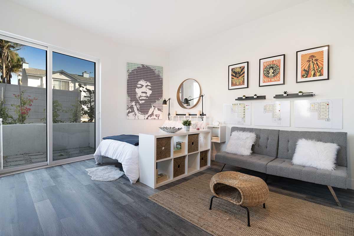 A modern bedroom featuring a gray sofa, a bed with white bedding, and a decorative area rug. The walls are adorned with framed art, including a portrait of a musician. Large windows allow natural light to fill the space, complementing the minimalist design and greenery visible outside.