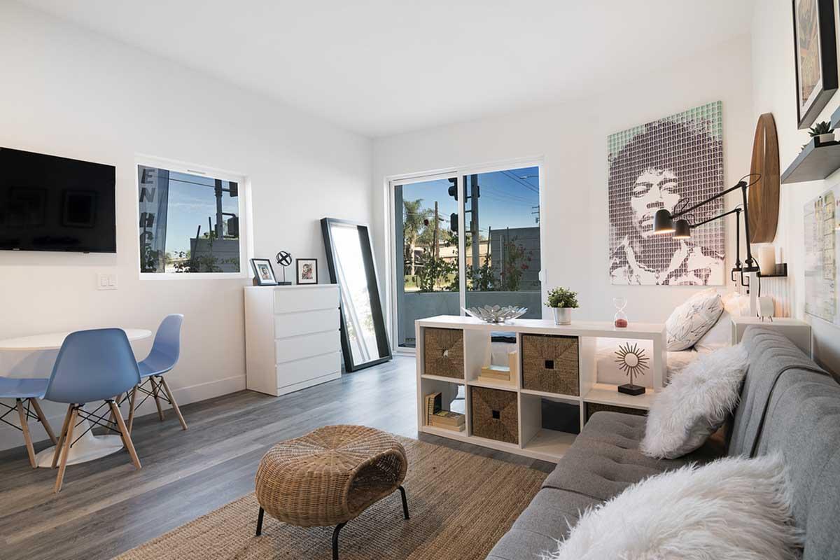 A cozy modern living space featuring a gray sofa with cushions, a round wicker ottoman, and a wooden shelf with decor items. There is a small dining table with blue chairs, a large mirror, and windows allowing natural light. A portrait is visible on one wall, adding character to the room.